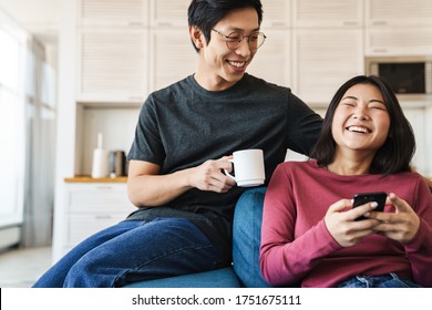 Cheerful Young Asian Couple Sitting On A Couch At Home, Woman Using Mobile Phone, Man Drinking Coffee