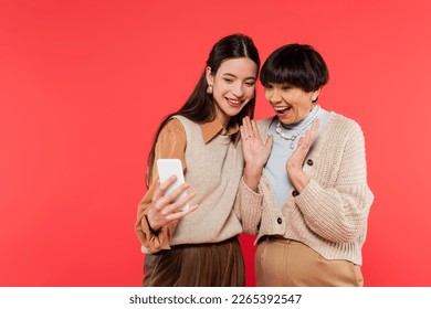 cheerful and young asian daughter holding smartphone near excited mother isolated on coral - Powered by Shutterstock