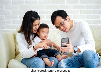 Cheerful Young Asian Family With Son Watching Video On Mobile Phone And Sitting On Sofa Couch At Home, Parents And Kid Enjoying Cartoon Movie