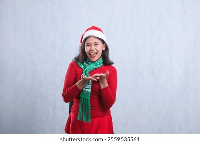 cheerful young asian female to camera both hands together presenting forward, wearing christmas sweater party outfit, santa hat and scarf, isolated on white background - Powered by Shutterstock