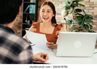 Cheerful young Asian female podcaster holding a questionnaire while interviewing male guest at workspace with mic and laptop