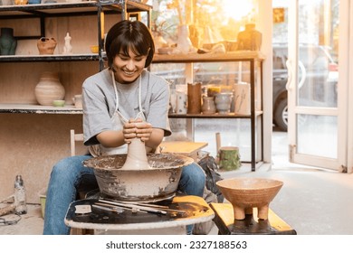 Cheerful young asian female potter in apron molding clay on pottery wheel near bowl and tools in ceramic studio at background at sunset, artisan creating unique pottery pieces - Powered by Shutterstock