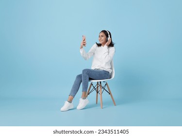 Cheerful young asian girl in wireless headphones sitting in armchair, listening to music, raising her arm, dancing to favorite song, enjoying popular soundtrack isolated on blue background. - Powered by Shutterstock