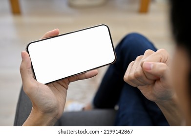 Cheerful Young Asian Guy Relaxing At Home, Watching Football Match Or Game Live Streaming On His Smartphone. Winning Gesture Expression. Phone White Screen Mockup. Cropped And Close-up Image