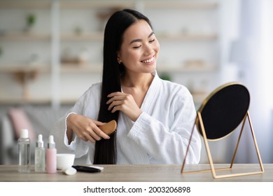 Cheerful young asian lady in bathrobe sitting in bedroom, combing her beautiful silky hair with wooden brush, looking at mirror and smiling, copy space. Hair care products concept