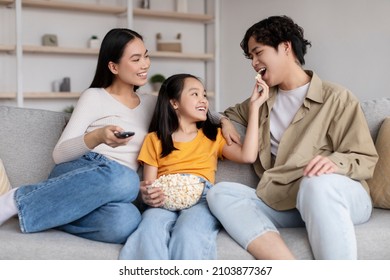 Cheerful Young Asian Lady, Man And Teenager Girl Watch Tv, Daughter Feeding Daddy Popcorn, Sits On Sofa In Living Room Interior. Happy Relationship, Family At Home, Technology And Movie Night Together