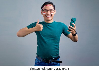 Cheerful young Asian man in casual t-shirt holding mobile phone and showing thumb up gesture isolated on grey background. indonesian independence day celebration concept - Shutterstock ID 2182268035