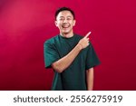 A cheerful young Asian man in a green T-shirt smiles widely while pointing to the side with one finger, standing confidently against a vibrant red background, exuding an inviting and positive vibe