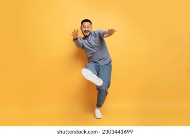 Cheerful young asian man having fun on yellow studio background, happy positive millennial guy reaching hands and showing shoe sole at camera, having fun, full length shot, copy space