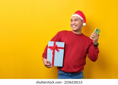 Cheerful young Asian man in Santa hat holding gift box with ribbon and mobile phone, looking away at copy space over yellow studio background. celebration Christmas holiday and New Year concept - Powered by Shutterstock