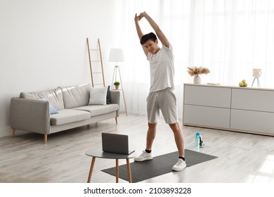 Cheerful young asian man stretching arm muscles, doing warmup exercises at home, looking at laptop screen. Smiling happy millennial guy having domestic training, standing working out indoors - Shutterstock ID 2103893228