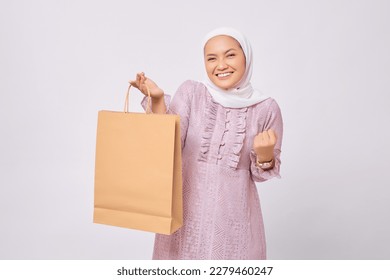 Cheerful young Asian Muslim woman wearing hijab and purple dress holding shopping bag and celebrating victory isolated on white studio background - Powered by Shutterstock