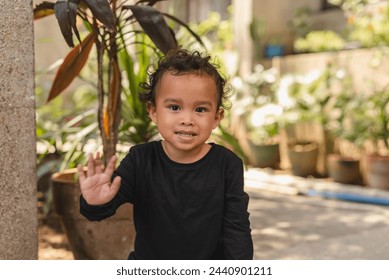 Cheerful young Asian toddler with curly hair waving his hand greeting warmly outdoors surrounded by greenery. - Powered by Shutterstock