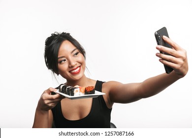 Cheerful Young Asian Woman Eating Sushi With Chopsticks Isolated Over White Background, Taking A Selfie