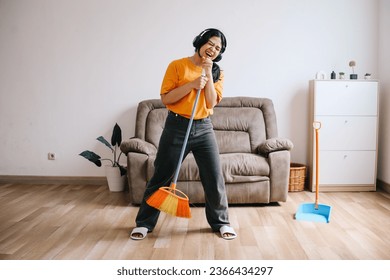 Cheerful young Asian woman in headphones using broom handle as microphone and having fun while cleaning house - Shutterstock ID 2366434297