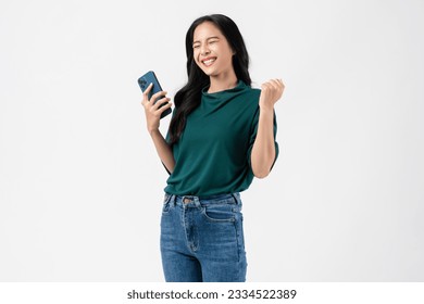 Cheerful young Asian woman holding smartphone with fists clenched celebrating victory expressing success on white background.