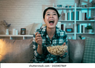 Cheerful Young Asian Woman With Popcorn Bowl Sitting On Sofa At Night Dark Home. Lady After Work Relax On Couch Laughing Joy With Open Mouth Wide Watching Tv Movie Funny Comedy Talk Show Program.