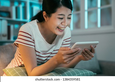 Cheerful Young Asian Woman Sitting On Couch Binge Watching Series Online On Tablet Pad In Dark Living Room Home. People Before Sleep Using Technology Concept. Laugh Housewife Enjoy Leisure Free Time