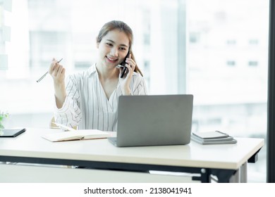 Cheerful Young Asian Woman Smiling And Looking Away While Having Smartphone Conversation In Office