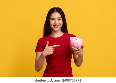 Cheerful young asian woman student pointing at pink piggybank in her hand, smiling at camera, isolated on yellow studio background. Savings, deposit, financial literacy, loan for education