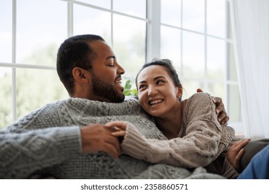 Cheerful young asian woman in sweater hugging and talking to african american boyfriend at home