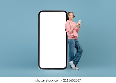 Cheerful Young Asian Woman With Wireless Headset, Smartphone Standing Next To Huge Mobile Phone With Empty Screen, Using Nice Entertaining Mobile App, Blue Background, Mockup, Panorama With Copy Space