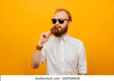 Cheerful Young Bearded Hipster Man Eating A Bar Of Chocolate Over Orange Background. Unhealthy Concept.