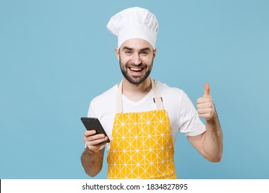 Cheerful young bearded male chef or cook baker man in apron white t-shirt toque chefs hat isolated on blue wall background studio portrait. Cooking food concept. Using mobile phone, showing thumb up - Shutterstock ID 1834827895