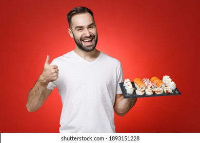 Cheerful Young Bearded Man 20s Wearing White T-shirt Showing Thumb Up Like Gesture Hold Makizushi Sushi Roll Served On Black Plate Traditional Japanese Food Isolated On Red Background Studio Portrait