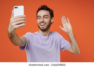 Cheerful young bearded man 20s in casual basic violet t-shirt standing doing selfie shot on mobile phone waving greeting with hand isolated on orange background studio portrait. Tattoo translate fun