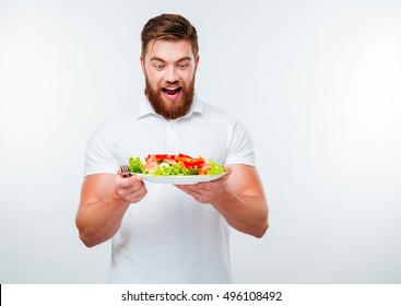 Cheerful young bearded man holding plate with fresh salad isolated on white background