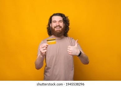 Cheerful Young Bearded Man Showing Thumb Up And His New Debit Card Over Yellow Background.