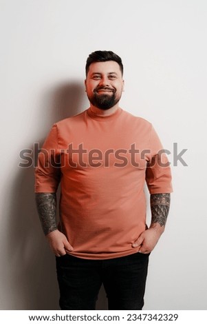 Cheerful young bearded plus sized male model in orange t shirt with tattooed arms looking at camera and smiling against white background in studio
