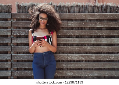 A cheerful young biracial female is leaning against a wooden fence with wattle inside, laughing, and holding the cell phone, with a copy space area on the right for text, your logo or advertising - Powered by Shutterstock