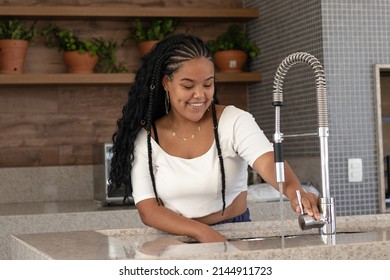 Cheerful Young Black Brazilian Woman With Braided Hair, White T-shirt, Earrings And Chain Opening The Faucet Of Her Kitchen Sink