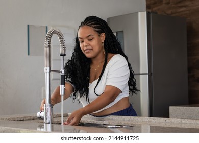 Cheerful Young Black Brazilian Woman With Braided Hair, White T-shirt, Earrings And Chain Opening The Faucet Of Her Kitchen Sink