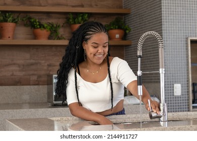 Cheerful Young Black Brazilian Woman With Braided Hair, White T-shirt, Earrings And Chain Opening The Faucet Of Her Kitchen Sink