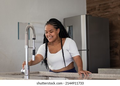 Cheerful Young Black Brazilian Woman With Braided Hair, White T-shirt, Earrings And Chain Opening The Faucet Of Her Kitchen Sink