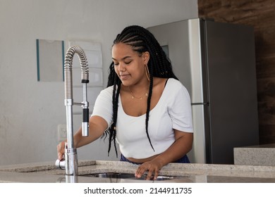 Cheerful Young Black Brazilian Woman With Braided Hair, White T-shirt, Earrings And Chain Opening The Faucet Of Her Kitchen Sink