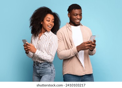 Cheerful young black couple back-to-back, deeply engaged in their smartphones, sharing light-hearted moment together against soothing light blue background, embodying modern communication - Powered by Shutterstock