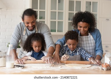 Cheerful Young Black Couple And Little Kids Baking Pie Together, Rolling Dough On Table With Flour And Ingredient Messy. Happy Parents Teaching Children To Cook Pastry, Having Fun In Kitchen