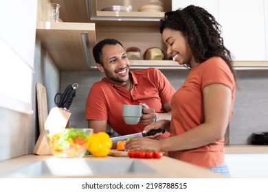 Cheerful Young Black Couple In Same T-shirts Talking, Wife Preparing Salad, Man Drink Coffee In Cozy Kitchen Interior. Cooking Together, Love And Relationship, Coffee Break And Healthy Food At Home