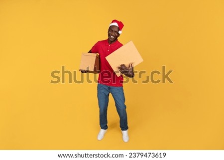 Cheerful young black delivery man in Santa Claus hat hold cardboard boxes, has fun, isolated on orange studio background. Order delivery service, work for Xmas, New Year