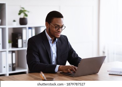 Cheerful young black entrepreneur in formal suit working with laptop, wearing glasses, office interior, copy space. Successful millennial african american businessman working on business project - Shutterstock ID 1879787527