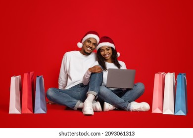 Cheerful Young Black Family In Santa Hats Sitting On Floor Among Colorful Shopping Bags, Using Laptop, Shopping On Internet While Winter Holidays, Christmas Sales Concept, Red Studio Background