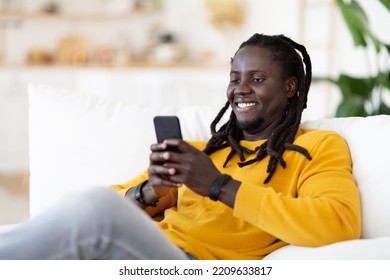 Cheerful Young Black Guy Messaging With Friends Or Browsing Internet On Smartphone At Home, Happy African American Man Using Mobile Phone While Relaxing On Comfortable Couch In Living Room