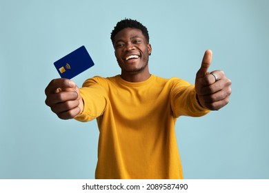 Cheerful young black guy in stylish yellow sweater showing thumb up and credit card, smiling at camera, posing alone on blue studio background, recommending his bank for online shopping or banking - Powered by Shutterstock