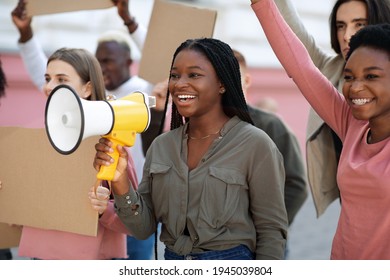 Cheerful young black lady leading international group of protestors, chanting slogans through megaphone. Active multiracial students with empty placards having strike or demonstration on the street