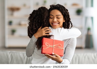 Cheerful Young Black Lady Mother With Gift Box Hugging Her Daughter, Celebrating Birthday Or Mothers Day Together At Home, Copy Space. Happy African American Mom And Kid Embracing