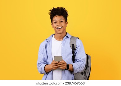 Cheerful young black male student with backpack comfortably using his smartphone, smiling at the camera standing against vivid yellow backdrop - Powered by Shutterstock